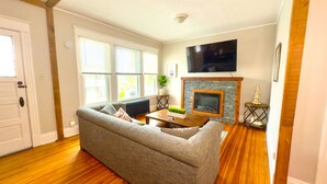 Una sala de estar con un sofá gris, piso de madera, una chimenea, un televisor montado y una puerta.