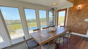 A wooden dining table with chairs, large windows, and a view of the outdoors. - Charming 6 bdr house-Chappaquiddick near Edgartown, beaches, 20x60pool,amenities (Edgartown)