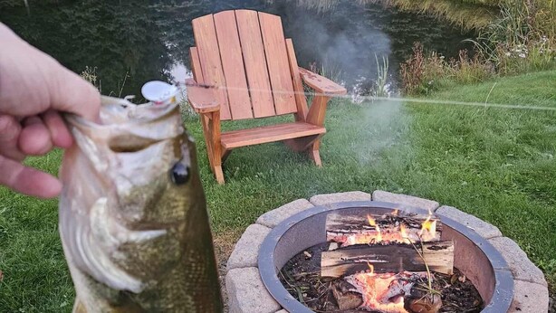 A person holding a fish near a stone fire pit with a wooden chair in the background.