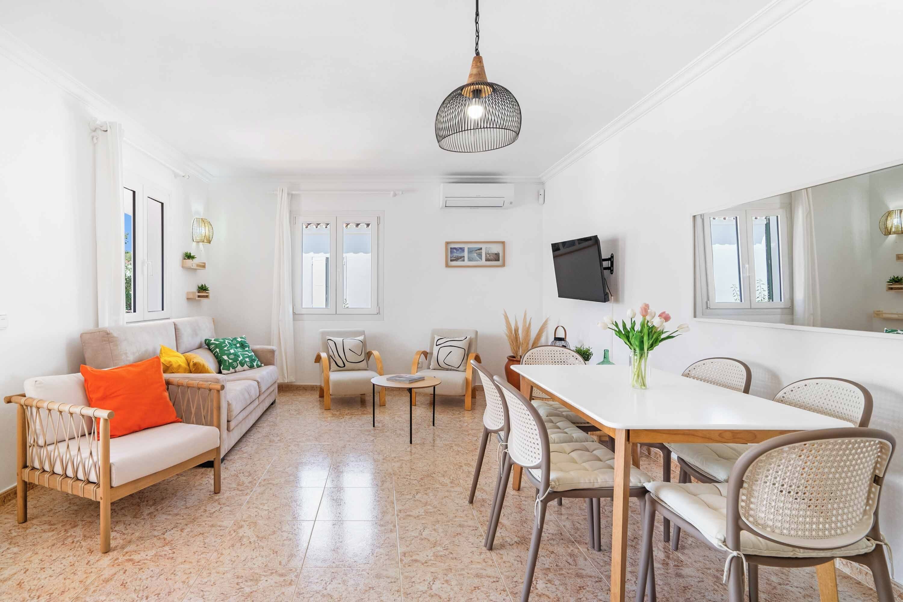 Un interior luminoso y blanco con un sofá, sillas, una mesa de comedor y una lámpara colgante.