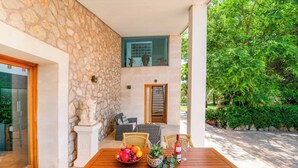 Un mur extérieur en pierre, une table en bois avec un panier de fruits et une bouteille de vin, des chaises en osier et une porte vitrée donnant sur un patio.