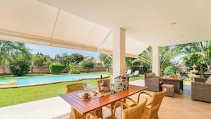 Une terrasse extérieure avec une table dressée pour le repas, entourée de chaises en osier, donnant sur une piscine et une végétation luxuriante.