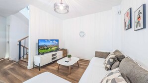A modern living room with a white sofa, a coffee table, and a TV on a white stand. There is a staircase leading to another room. - Villa Julaga (La Oliva)
