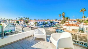 Un balcón con dos sillas de plástico blanco, una mesa de vidrio y vistas a una zona residencial con palmeras y casas.