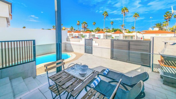 A rooftop pool area with a glass balustrade, a table with chairs, and a view of palm trees and buildings in the background. - Villa Julaga (La Oliva)