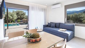 A modern living room with a glass dining table, a blue sofa, and a view of a pool and trees. - Villa Averto Anna (Lefkada)