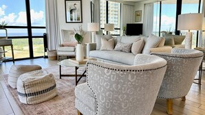 Ein geräumiges Wohnzimmer mit einer großen Couch, zwei Sesseln, einem Couchtisch und Meerblick durch große Fenster.