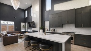 A modern kitchen with dark cabinets, a large island with stools, a flat-screen TV, and a chandelier. - Downtown Dallas Home  (Dallas)