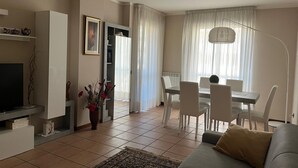 A living room with a sofa, a dining table with chairs, a TV, and a vase with flowers.