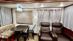 A compact RV interior with a sofa, dining table, two chairs, and a single reclining chair.