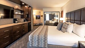 Quarto de hotel moderno com uma cama grande, escrivaninha embutida, cozinha compacta e banheiro visível por uma porta aberta.