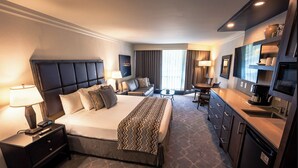 Quarto de hotel com uma cama grande, uma mesa com cafeteira, uma televisão e uma área de estar com mesa e cadeiras.