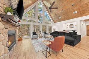 A spacious living area with a stone fireplace, large windows, a ceiling fan, and a cozy seating arrangement.