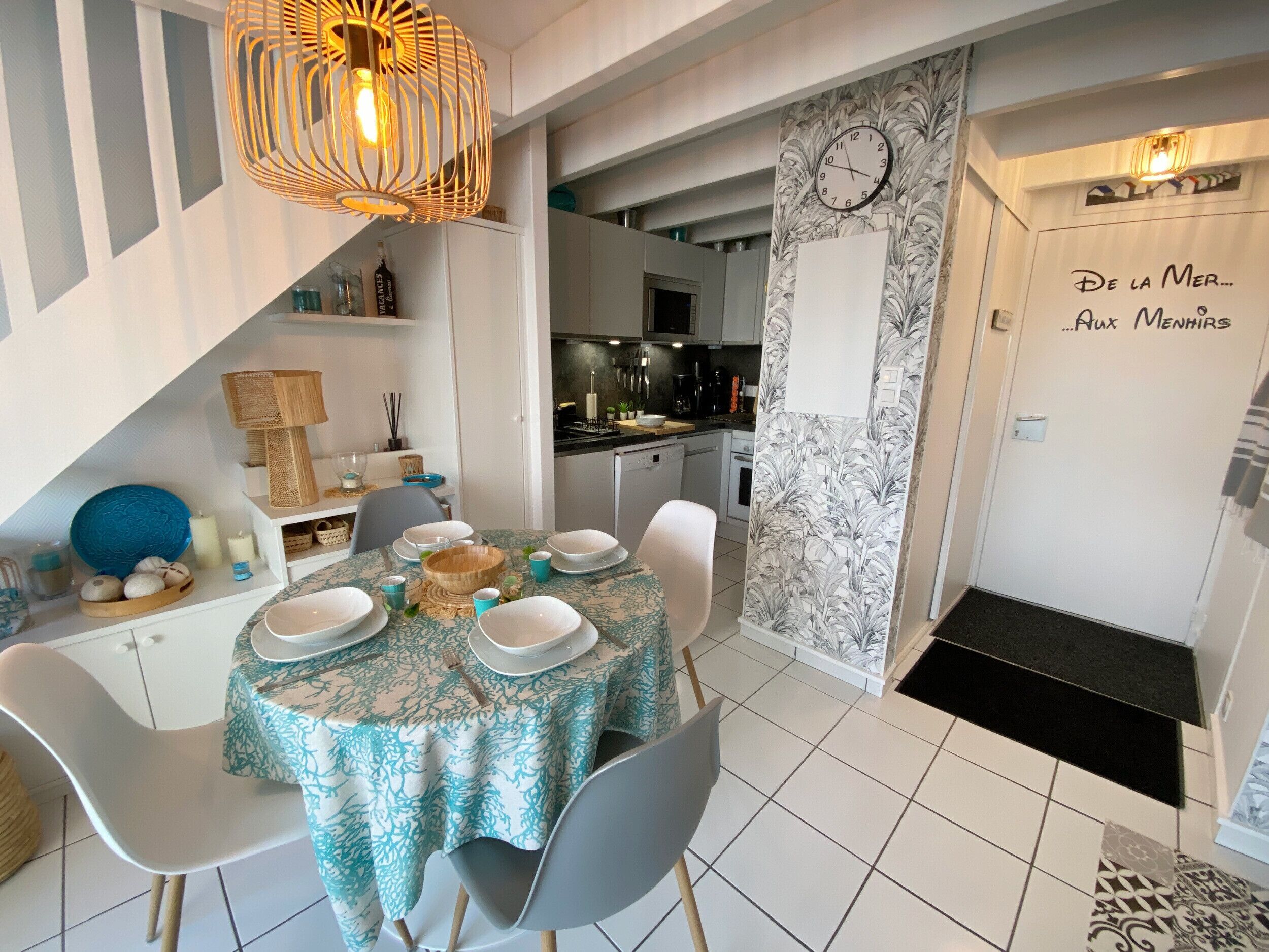 A dining area with a table set for four, chairs, a decorative hanging light, and a kitchen in the background.