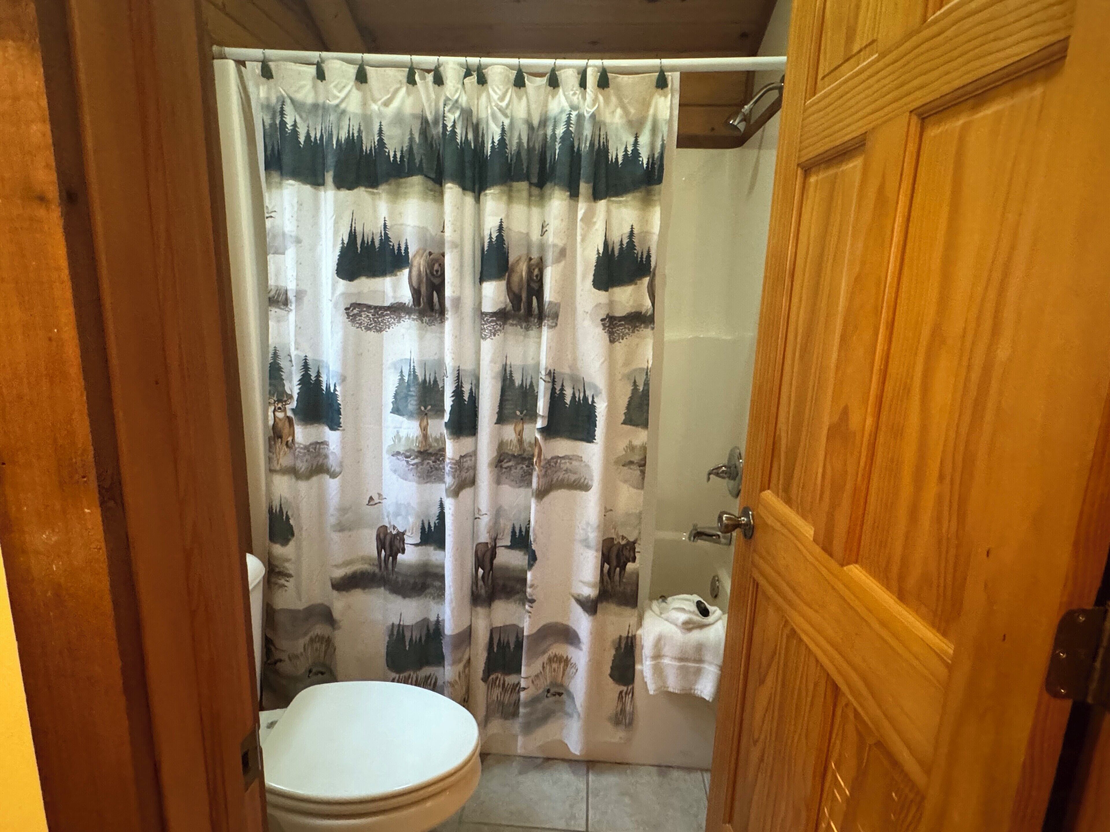 A bathroom with a shower curtain featuring forest and wildlife prints, a wooden door, and a white toilet.