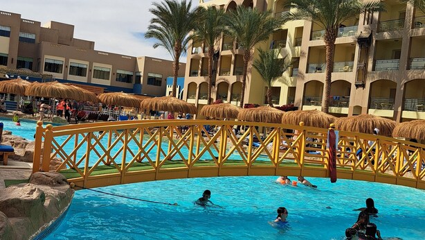 Un área de piscina con un puente de madera, palmeras y un edificio de varios pisos con balcones.