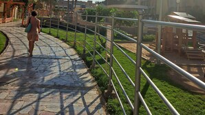 Un camino pavimentado con barandilla metálica, palmeras y una zona de juegos.