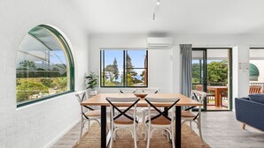 Een moderne eetruimte met een houten tafel en stoelen, grote ramen met uitzicht op bomen en een wateroppervlak, en een open indeling die leidt naar een balkon met buitenmeubilair.