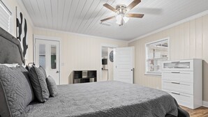Une chambre avec un grand lit, un ventilateur de plafond, une commode et une fenêtre avec des stores.