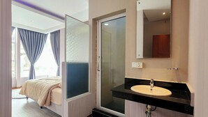 Habitación de hotel con cama, baño con lavamanos y puerta de cristal que da a la ducha.