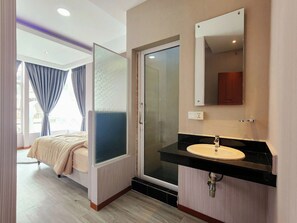 A hotel room with a bed, a bathroom with a sink, and a glass door leading to a shower.