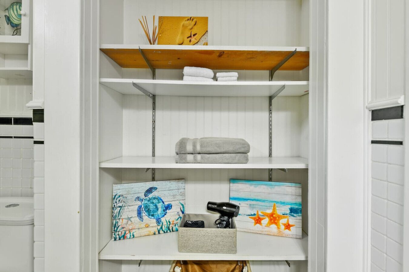 A bathroom with white tiled walls, a white sink, and a shelf with towels and decorative items.