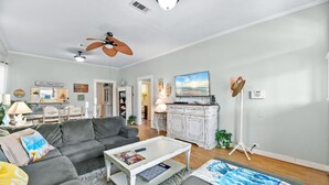 Ein Wohnzimmer mit grauem Sofa, weißem Couchtisch und einem Fernseher. Es gibt einen Deckenventilator, eine Lampe und eine Pflanze.