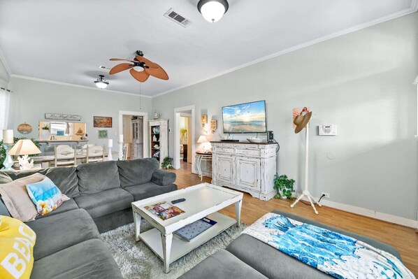 A living room with a grey sofa, white coffee table, and a television. There is a ceiling fan, a lamp, and a plant.