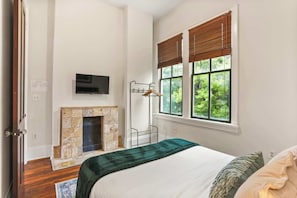 A bedroom with a fireplace, a bed with a green blanket, a TV mounted on the wall, and a window with blinds.