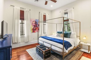 A four-poster bed with a blue blanket, a wooden floor, a desk with a chair, and a suitcase.
