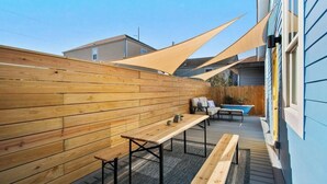 Eine Holzterrasse mit Bänken und einem Tisch, ein Schwimmbecken und ein blaues Gebäude im Hintergrund.