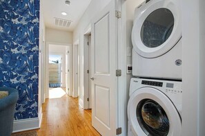 Une buanderie équipée de machines à laver et de sèche-linge superposés, avec un plancher en bois et une porte donnant sur une autre pièce.