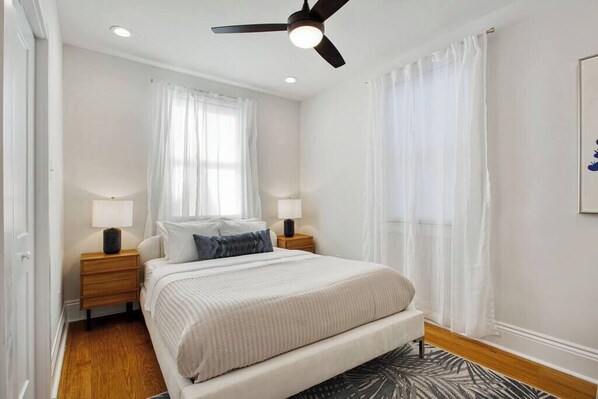 Un dormitorio con una cama, dos mesitas de noche, un ventilador de techo y una ventana con cortinas de gasa.
