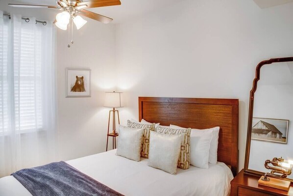A hotel room with a bed, a ceiling fan, a bedside lamp, a mirror, and a framed picture on the wall. - NOLA Sanctuary Summar (New Orleans)