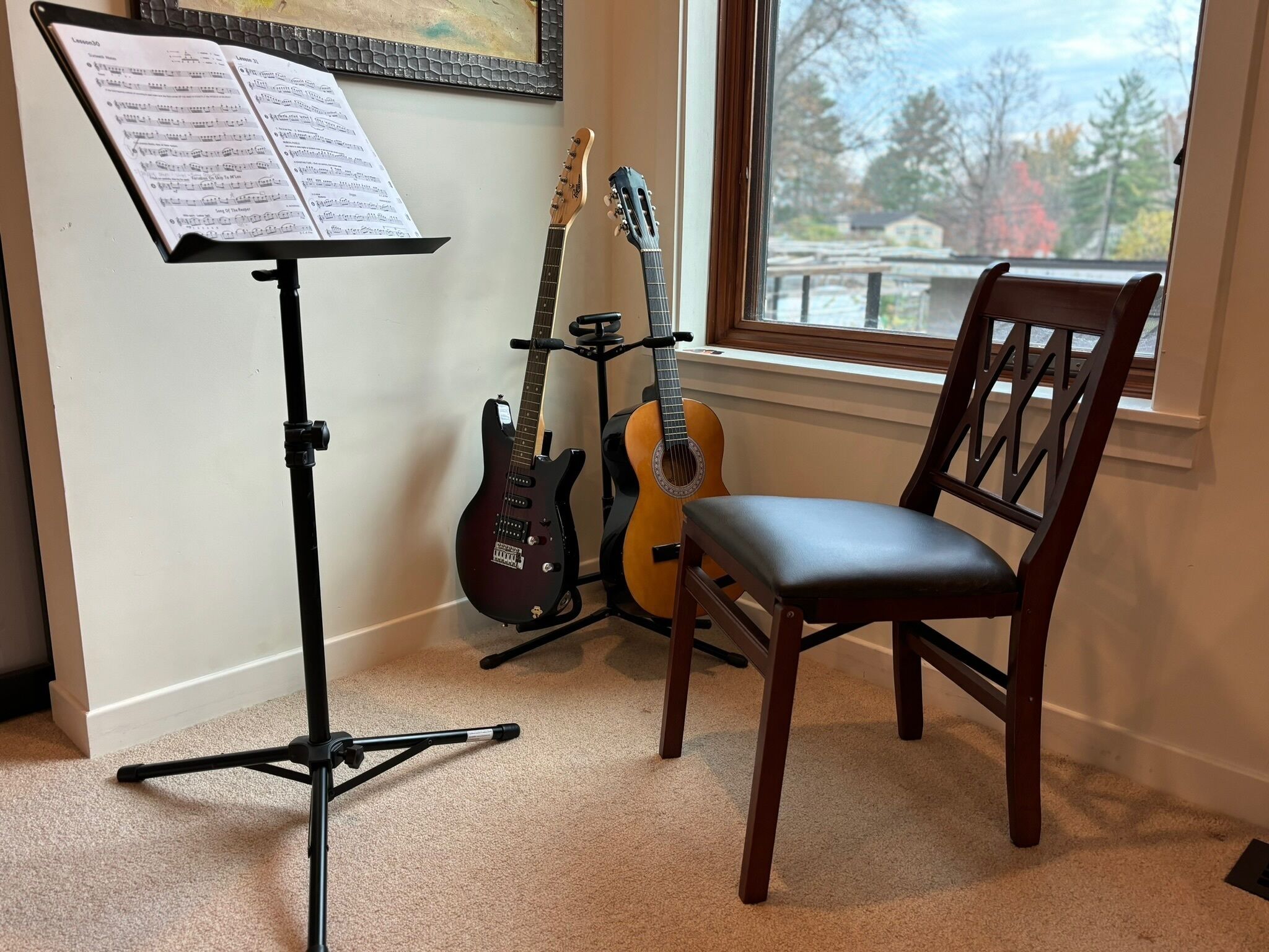 A music stand with sheet music, two guitars, and a chair in a room with a window.