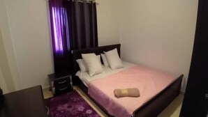 Un dormitorio con una cama, una mesita de noche, un tocador, un tapete floral morado y una ventana con cortinas moradas.