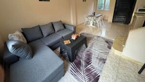 Una sala de estar con un sofá gris, una mesa de centro y una mesa de comedor con sillas. Se ve una ventana y una puerta al fondo.