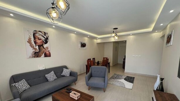 A modern living room with a grey sofa, a blue armchair, and a wooden coffee table. There is a dining area in the background with a table and chairs. - 903 . Spacious 3BR Apt., 2 Baths, Gated Community (Al Sheikh Zayed)