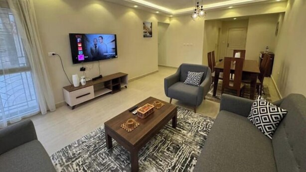 A modern living room with a flat-screen TV, a coffee table, a sofa, and a dining area. - 803 . Spacious 3BR Apt., 2 Baths, Gated Community (Al Sheikh Zayed)