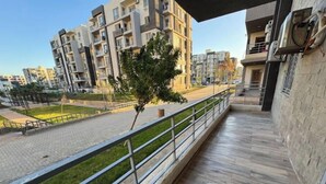 A balcony with a view of modern apartment buildings, a paved walkway, and a small tree. - 803 . Spacious 3BR Apt., 2 Baths, Gated Community (Al Sheikh Zayed)