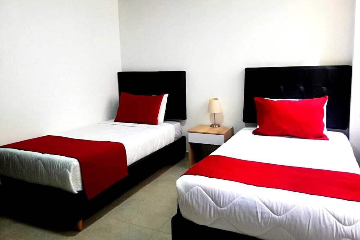 Una habitación de hotel con dos camas individuales, cada una con cubrecama y almohadas rojas y blancas. Entre las camas, una mesita de noche con lámpara.