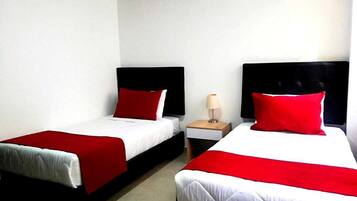 A hotel room with two single beds, each with a red and white bedspread and matching pillows. There is a small bedside table with a lamp between the beds.