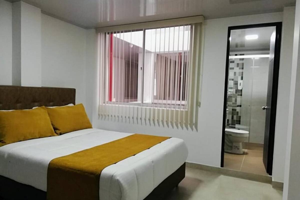 Un dormitorio con cama, ventana con persianas y baño visible a través de una puerta abierta.