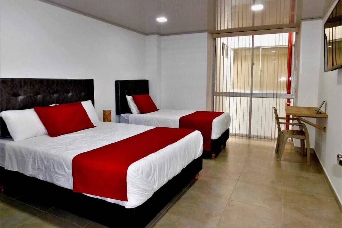Habitación de hotel con dos camas, cada una con ropa blanca y detalles rojos, un escritorio de madera y un ventanal con persianas.