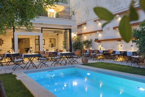Uma área de piscina ao ar livre com assentos e mesas, cercada por vegetação e um edifício com fachada de pedra.