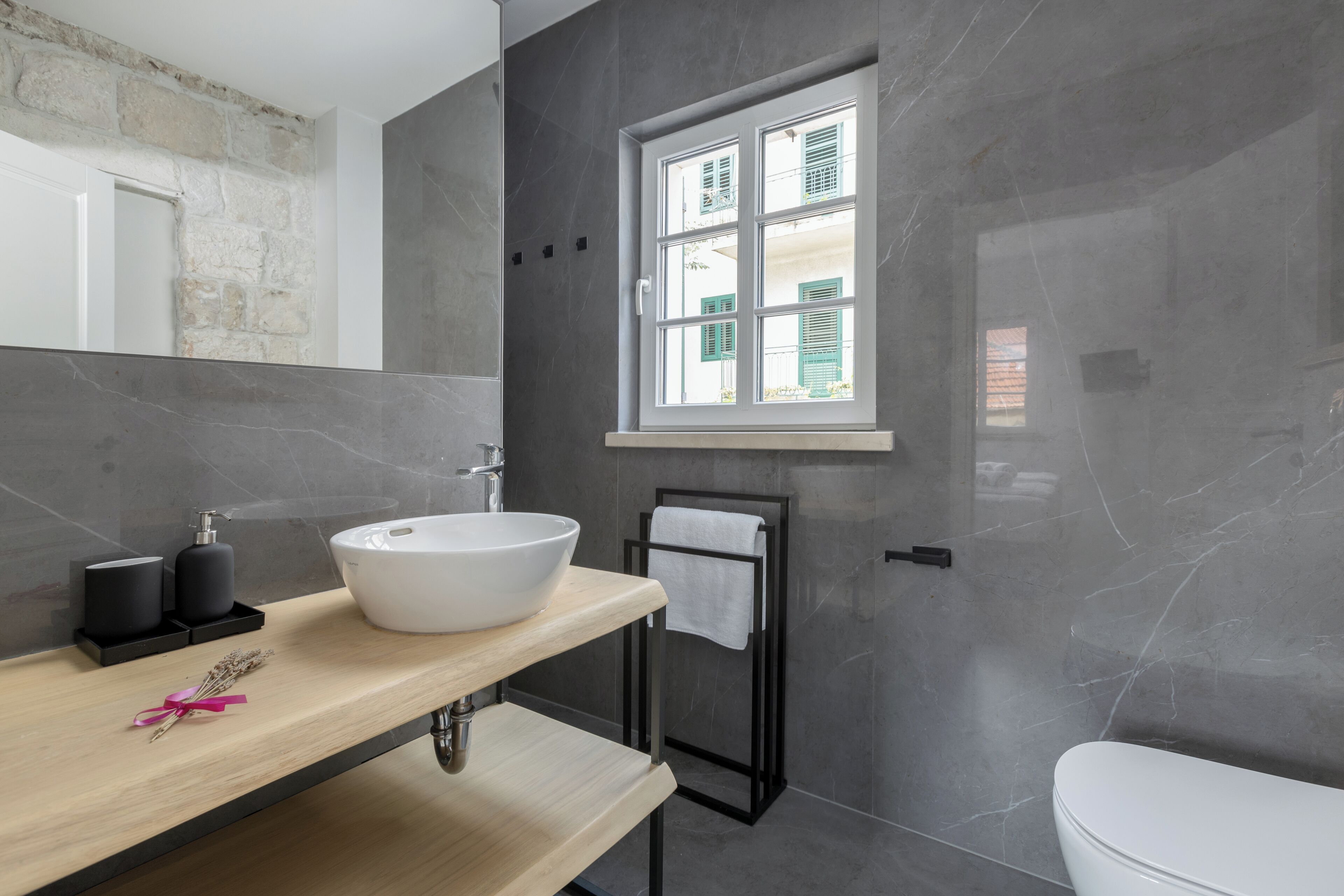 Un baño moderno con un lavamanos grande de color blanco, un mueble de tocador de madera, un perchero para toallas y una ventana con contraventanas.