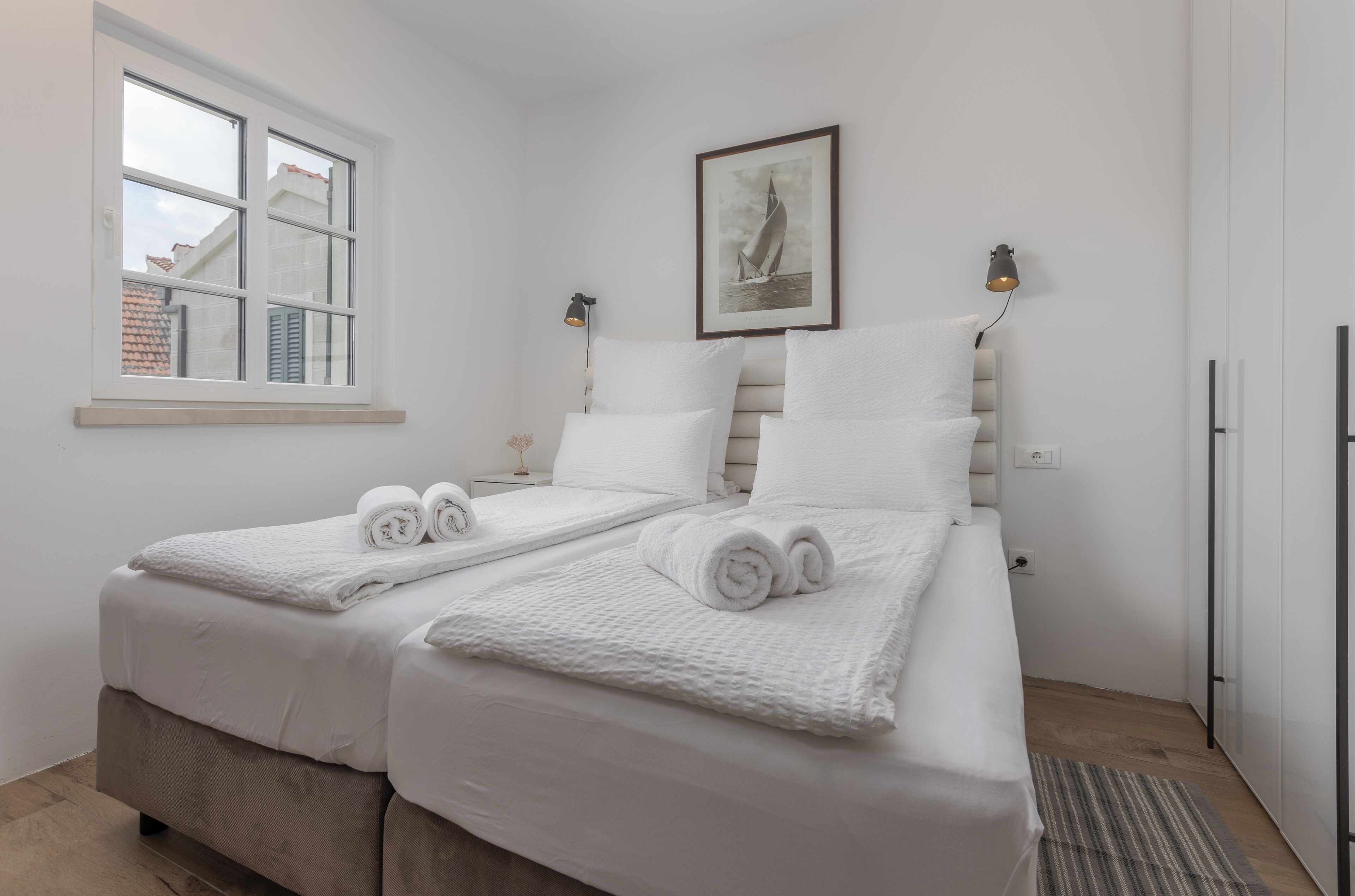 A bedroom with two beds, white linens, a framed picture on the wall, and a window with a view of buildings.