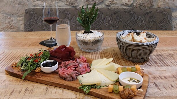 A wooden table with a charcuterie board, a glass of red wine, a glass of water, a potted plant, and a basket of bread. - Casa Columba (Makarska)