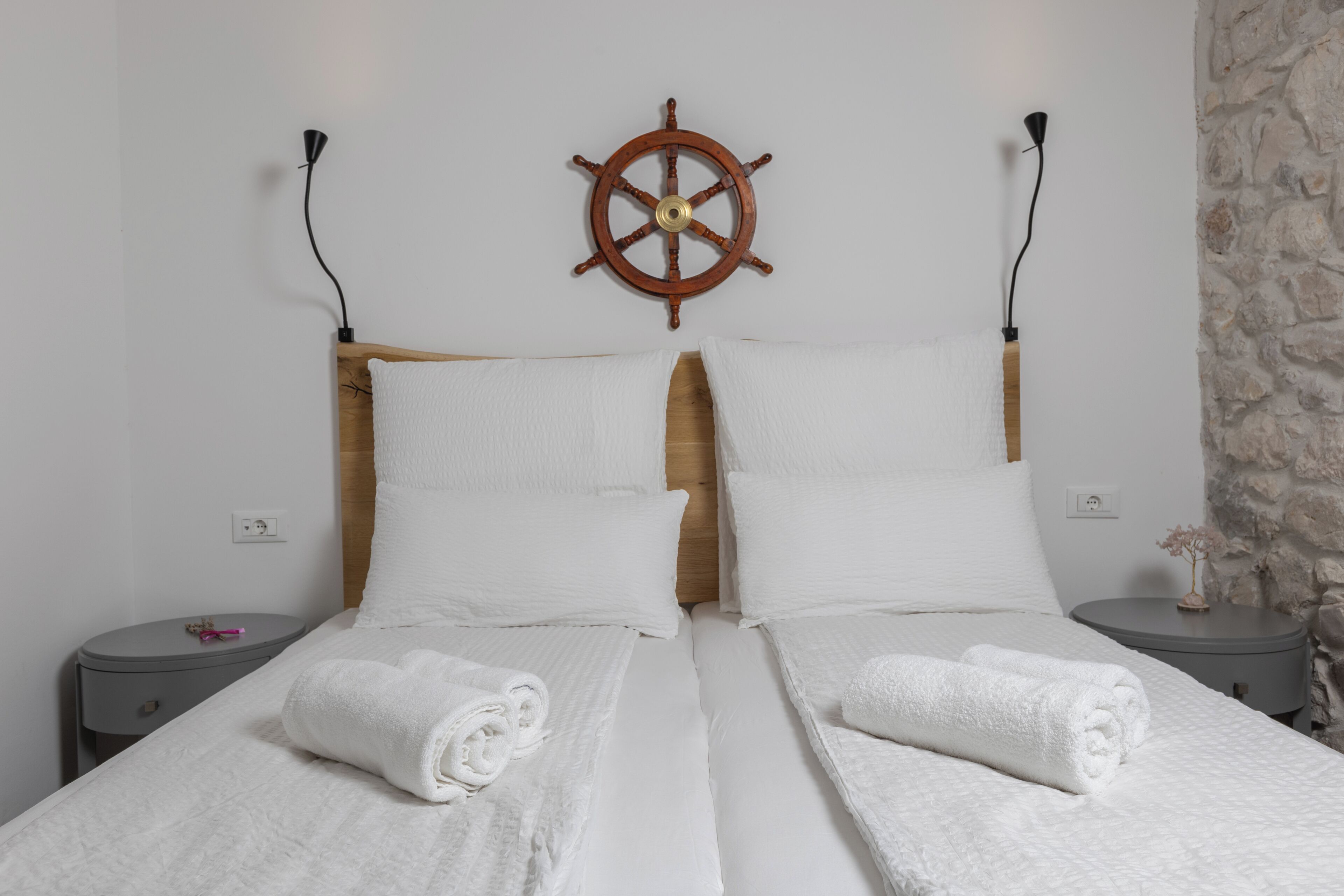 Una cama doble con sábanas blancas y dos almohadas, un cabecero de madera, un timón de barco montado en la pared y dos mesitas de noche con lámparas.