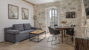 A modern living room with a grey sofa, a wooden coffee table, and a stone wall. There are two framed pictures on the wall, a dining table with chairs, and a large window. - Casa Columba (Makarska)
